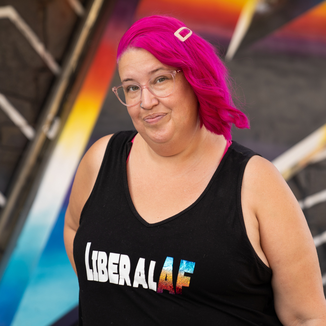 Meg stands confidently with a smirk, with hot pink hair and "LiberalAF" on her tank top