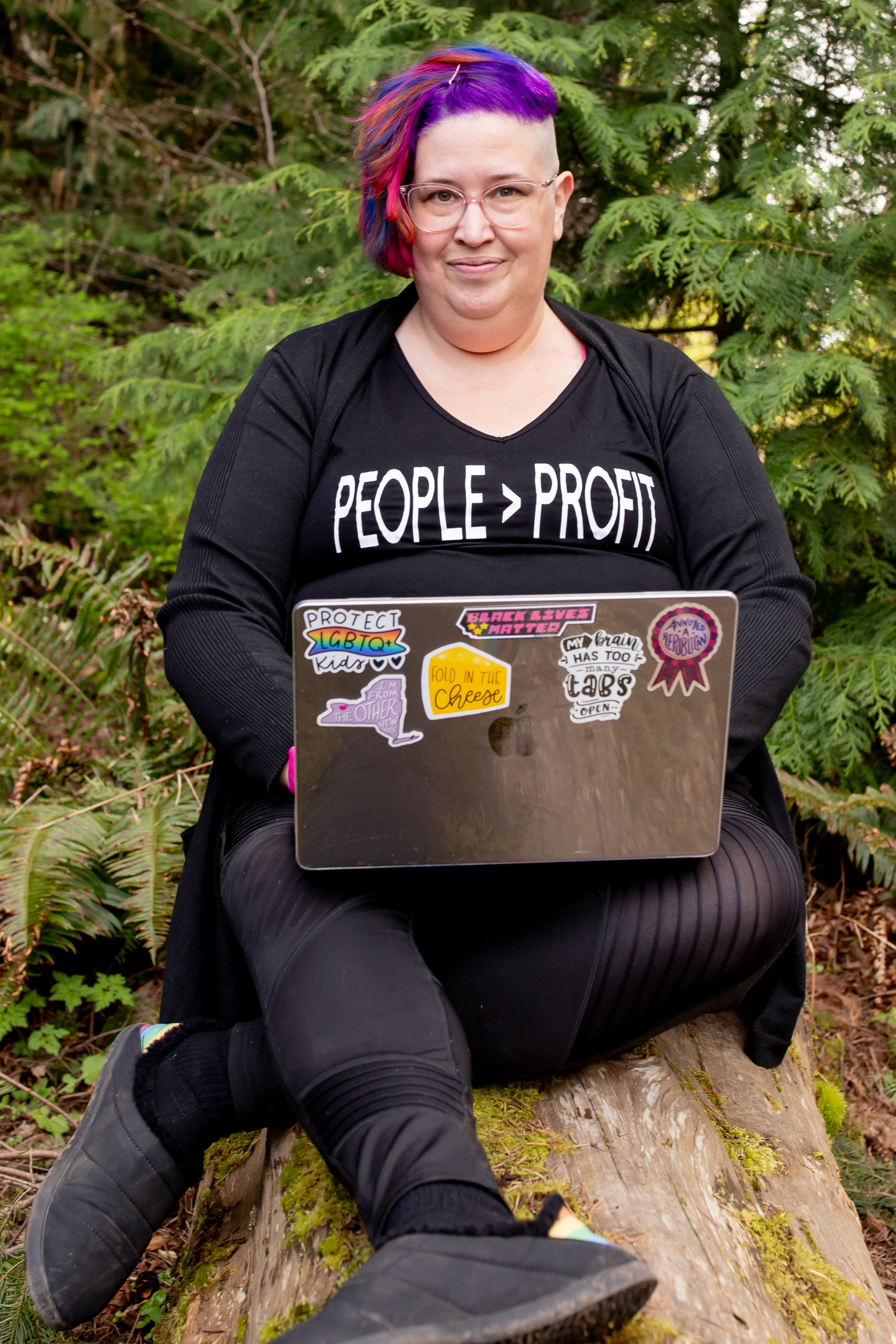Meg sits on a log in the woods working on her laptop. Her hair is purple and pink and her shirt says "people > profits"